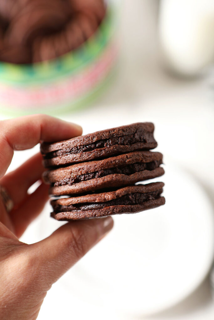 A hand holds three chocolate sandwich cookies with mint chocolate ganache over a plate and a cookie tin