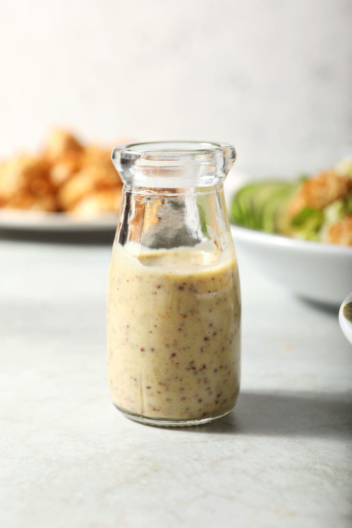 A small jar of Creamy Honey Mustard Dressing on a green surface in front of bowls of salad and chicken nuggets