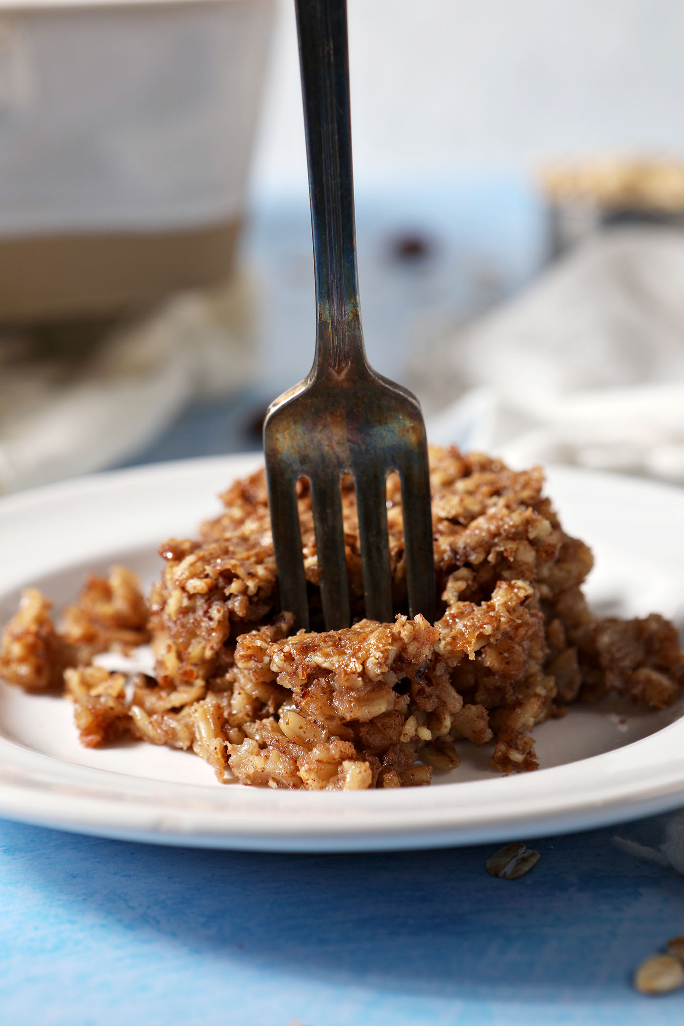 A fork cuts into a baked oatmeal bar on a white plate sitting on a blue surface near white linens and a casserole dish