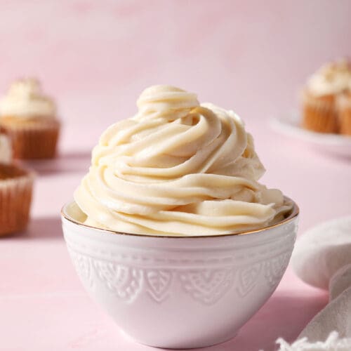 a bowl of cream cheese frosting on a pink surface in front of cupcakes