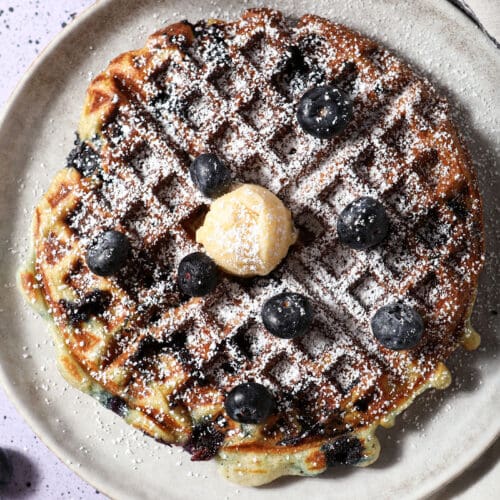 A blueberry waffle topped with powdered sugar, fresh blueberries and a dollop of honey butter on a gray plate on a purple speckled surface