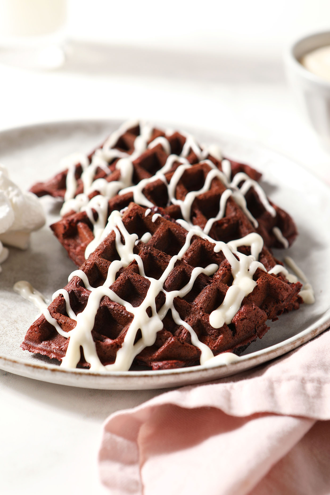 A grey plate of red velvet waffles drizzled with cream cheese icing on a white surface next to a pink linen