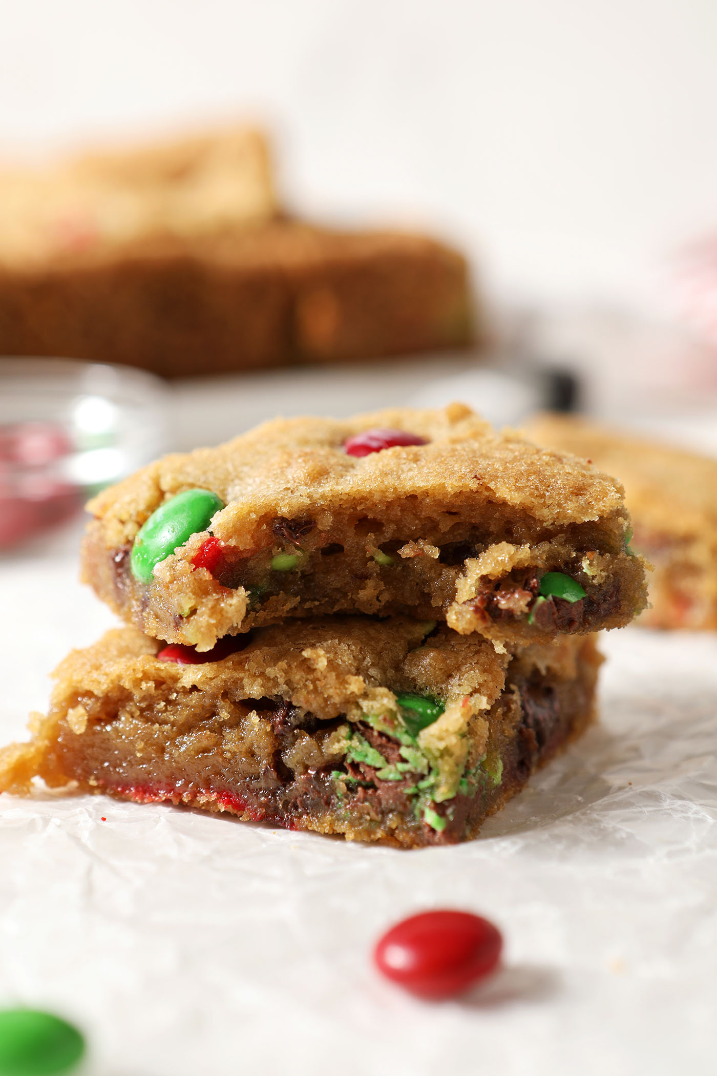Close up of stacked Christmas M&M Cookie Bars, one bitten into, on crinkled white parchment paper